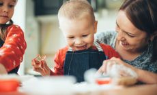 happy baby cuts pastry dough as mother and sibling look on